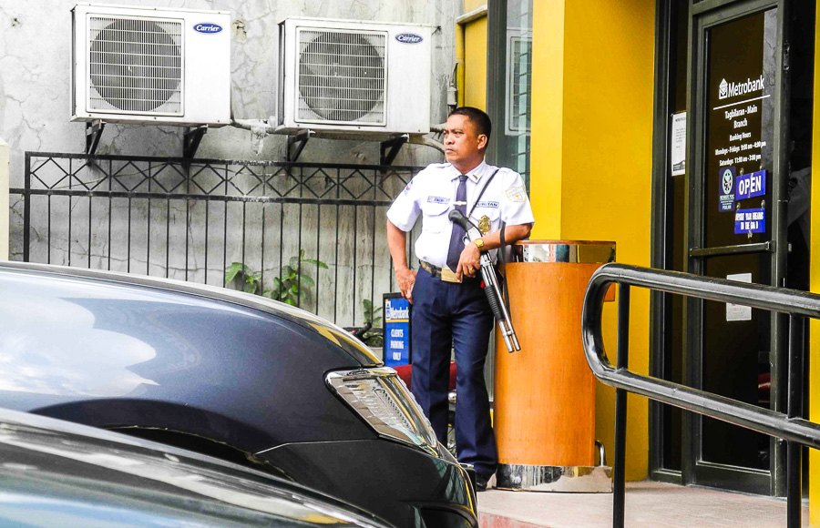 A stern security guard at the bank entrance never lets his weapon out of his hands
